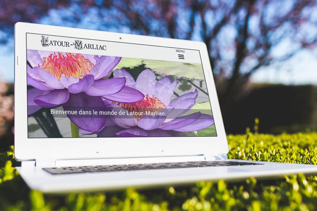 Un ordinateur portable affiche la page d'accueil du site latour-marliac.com avec une image de nénuphars violets en arrière-plan, posé sur de l'herbe dans un cadre extérieur ensoleillé.